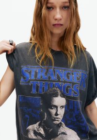 Tricou unisex grafic din material negru uzat, cu croială supradimensionată, având text albastru "STRANGER THINGS" și imprimeu central cu un personaj.