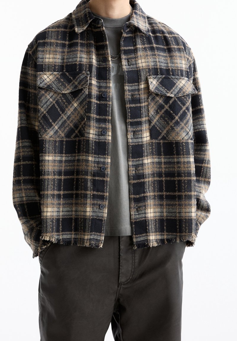 Homme portant une chemise à carreaux beige et noire par-dessus un t-shirt gris avec graphique, assortie de pantalons sombres et d'un collier en argent.