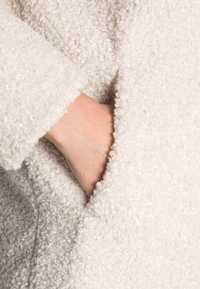 Cream-colored coat with a soft, textured fabric. Features a side pocket and loose-fitting sleeves.