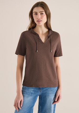 Young woman wearing a brown short-sleeve top with a V-neck and drawstrings, paired with blue jeans, standing against a plain background.