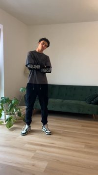 Mustat cargo-housut ja harmaa pitkähihainen paita, jossa on graafisia kuvioita. Mustat ja valkoiset lenkkarit. Taustalla vihreä pehmustettu sohva.