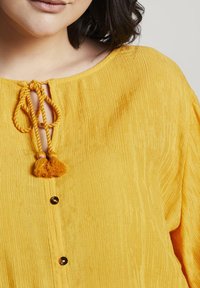 Top giallo con texture, dotato di scollatura rotonda, caratterizzato da una chiusura con fiocco e nappine e bottoni scuri decorativi sul davanti.