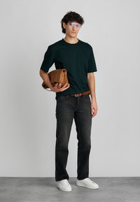 Mörkgrön t-shirt med korta ärmar, mörka jeans, vita sneakers, håller i en tanfärgad läderväska, kontrasterande brunt bälte och glasögon med ljus båge.