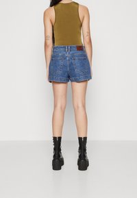Džínové šortky s vysokým pasem, modré barvy a roztřepeným lemem, doplněné olivově zeleným tílkem a černými kotníkovými botami na šněrování.