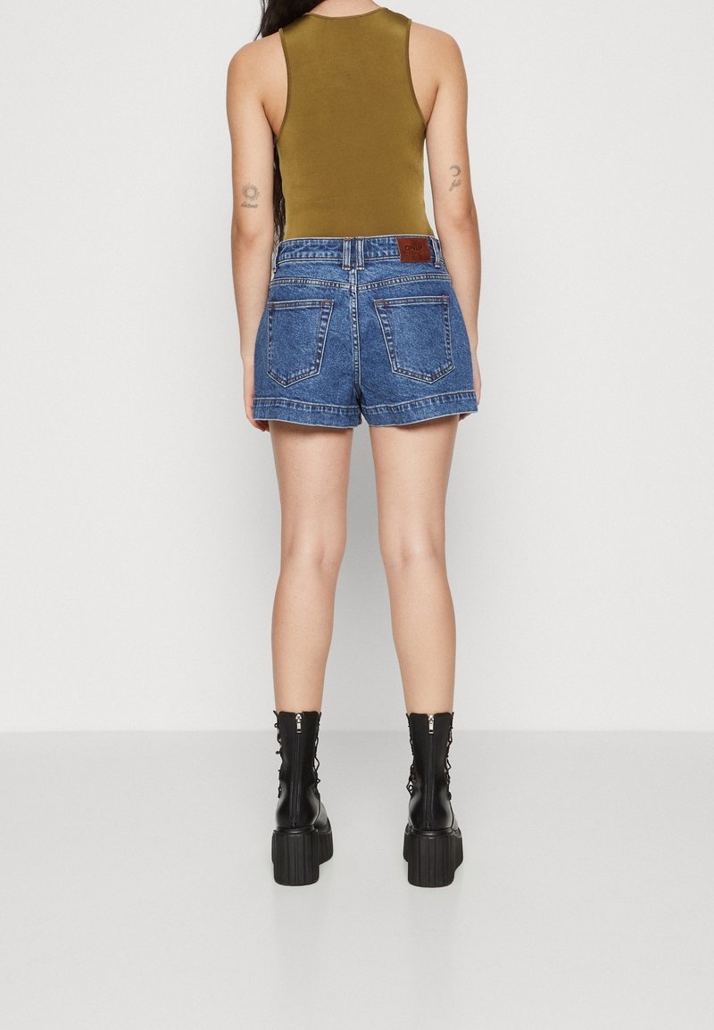Džínové šortky s vysokým pasem, modré barvy a roztřepeným lemem, doplněné olivově zeleným tílkem a černými kotníkovými botami na šněrování.