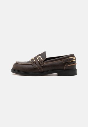 Mocasin de cuero marrón oscuro con dobles hebillas doradas y un tacón bajo negro sobre un fondo blanco.