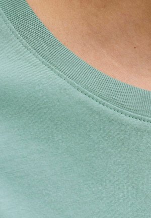 Roheline heledam, soonikestega crew neck T-särk, mis on seljas palja naha peal, kaelaääre õmblused on nähtavad.