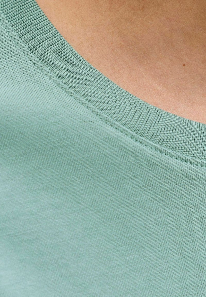 Close-up van een lichtgroen, geribd T-shirt met ronde hals, gedragen op blote huid met zichtbare stiksels langs de halslijn.