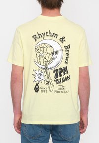 Camiseta de algodón amarilla con gráfico impreso que presenta una luna tocando el saxofón, el texto "Rhythm & Brews" y detalles en negro.