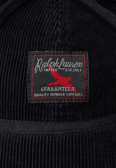 Svart keps i manchester med en rektangulär lapp. Lappen har vit och röd text, en röd fågellogo och texten "Guaranteed Quality Outdoor Supplies."