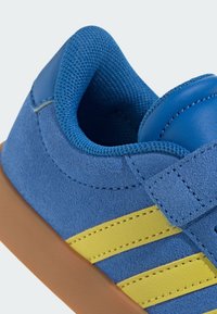 Baskets en suede bleues avec des rayures jaunes, doublure en maille texturée et semelle en caoutchouc marron. Équipées d'une sangle Velcro pour un maintien sécurisé.