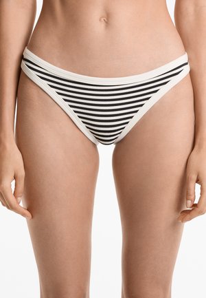 Kvinna som bär en bikini underdel med svartvita horisontella ränder, stående med armarna avslappnat vid sidorna mot en vit bakgrund.
