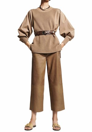 Kobieta ubrana w beżowy sweter z bufiastymi rękawami, z brązowym paskiem, szerokie brązowe skórzane spodnie, złoty naszyjnik z łańcuszkiem oraz plecione sandały z klamrą.