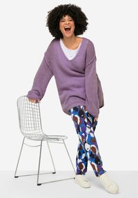 Ampio maglione lavorato a maglia di colore viola con scollo a V profondo, abbinato a leggins colorati con fantasie che presentano volti astratti. Sneakers bianche e una sedia in metallo.