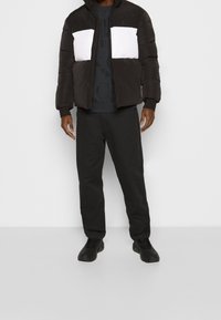 Hombre vestido con una chaqueta acolchada negra y blanca, pantalones negros y zapatillas negras, de pie contra un fondo blanco liso.