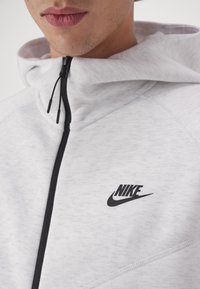 Světle šedá mikina na zip s černým logem Nike. Má vysoký límec, texturovanou tkaninu a kontrastní černý zip.
