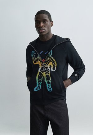 Junger Mann trägt schwarzen Kapuzenpullover mit Reißverschluss und farbenfroher Astronautengrafik, Hände in den Taschen, steht vor einem schlichten hellen Hintergrund.