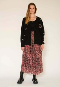 Cardigan noir avec des patchs brodés, superposé sur une jupe florale rose et noire. La tenue est accompagnée de bottes noires jusqu'aux genoux.