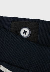 Marineblauer gerippter Stoff mit einem schwarzen Textilakzent. Verfügt über einen transparenten Knopf mit vier Löchern. Fokus auf dem Kragen-Detail und der Naht.