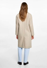 Béžový vlněný kabát s rovným střihem, dlouhými rukávy a rozparkem na zádech. Kombinován se světle modrými džíny a černými botami, má hladkou texturu.