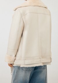 Krémově zbarvený kabát z ovčí vlny s měkkým, texturovaným límcem a manžetami, s rafinovaným střihem a viditelnými detaily švů.