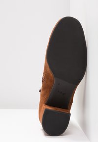 Chaussure en daim marron avec une semelle en caoutchouc texturé. Présente un talon modéré, des coutures sur les côtés et un nœud décoratif près de l'ouverture.