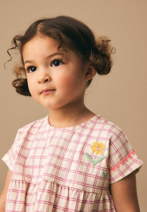Junges Mädchen mit lockigem Haar in zwei Dutts trägt ein rosa-weiß kariertes Kleid, das mit einer gelb bestickten Blume verziert ist.