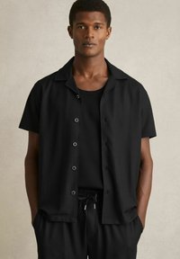 Cămașă neagră cu mânecă scurtă, cu textură ribbată, cu deschidere cu nasturi în față și guler plat, purtată peste un top negru fără mâneci și pantaloni asortați.