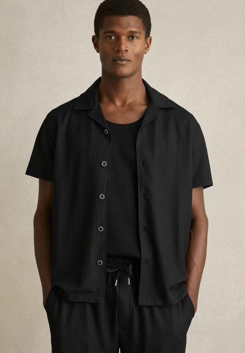Camicia nera a maniche corte con una texture a costine, davanti con bottoni e colletto piatto, indossata sopra un top senza maniche nero e pantaloni abbinati.