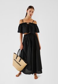 Fekete, vállról lelógó crop top és hozzá illő hosszú szoknya, gumírozott derékrésszel. A modell egy fekete részletekkel díszített szalmakosarat tart.