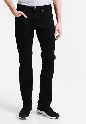 Man draagt zwarte slim-fit jeans en zwarte sneakers met witte zolen, staand met één hand in zijn zak tegen een witte achtergrond.
