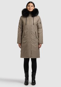 Beež pikk mantel musta kunstkarusnahast kapuutsiga, esikinnis lukuga, kahe küljetasku ja stiilse disainiga. Kandmisel mustade retuuside ja pahkluu-saabastega.