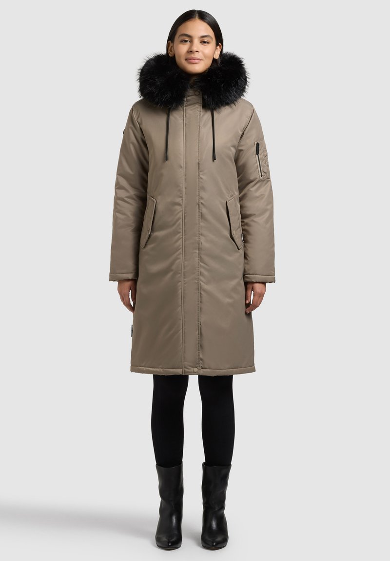 Beež pikk mantel musta kunstkarusnahast kapuutsiga, esikinnis lukuga, kahe küljetasku ja stiilse disainiga. Kandmisel mustade retuuside ja pahkluu-saabastega.