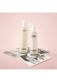 Twee Babor huidverzorgingsflessen op een roze achtergrond: een romige witte reiniger en een hydraterende booster, beide met pompflacons en minimale zwarte tekst.
