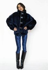 Marineblå faux pelsjakke med stor krave og knapper foran; kombineret med skinnende blå velourleggings og knælange sorte støvler.