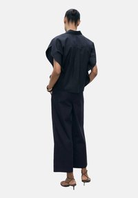 Zwarte cropped top met korte mouwen en een rugplooien, gecombineerd met losse, donkere broek en sandalen met hakken en bandjes.