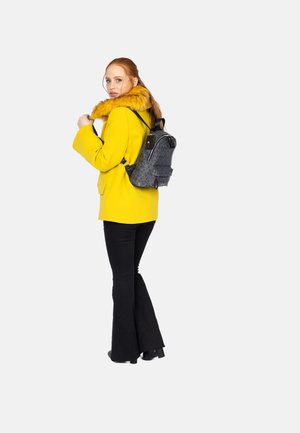 Mochila negra con estampado y correas ajustables, combinada con un abrigo amarillo brillante que presenta un cuello de piel sintética. Modelo con pantalones de campana negros.