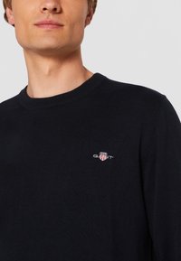 Navy knit sweater with a round neck, featuring a small embroidered logo on the chest. Smooth texture, fitted design, and long sleeves.