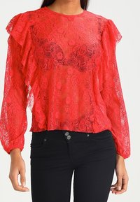 Top de encaje rojo con mangas acampanadas, textura transparente y cuello sólido. Combinado con pantalones negros ajustados. Patrón floral notable visible.