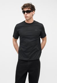 Νεαρός άντρας που φοράει μαύρο T-shirt Timberland, μαύρο παντελόνι και μαύρα γυαλιά ηλίου, στέκεται με τα χέρια πίσω από την πλάτη μπροστά σε ένα λευκό φόντο.