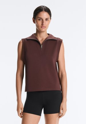 Kvinde iført en ærmeløs brun lynlåstop og sorte shorts, stående mod en ensfarvet lys baggrund.