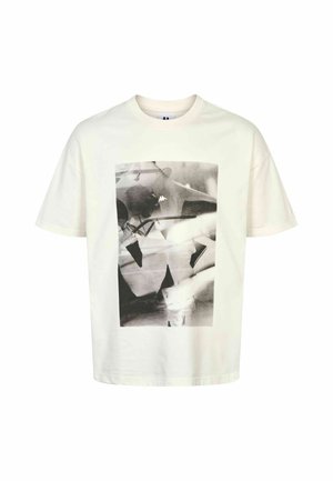 Ung mand med tatoverede arme iført hvid t-shirt med sort og hvidt abstrakt tryk, trækker let i t-shirten fremad, mod ensfarvet baggrund.