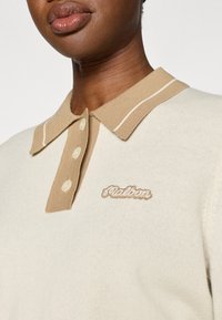 Κοντινό πλάνο ατόμου που φοράει κρεμ πλεκτό πόλο πουλόβερ με μπεζ γιακά, τρία κουμπιά και κεντημένο κείμενο "Galdin" στο στήθος.