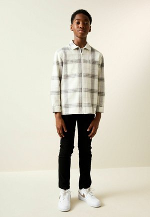 Adolescent în picioare lângă un perete simplu, purtând o cămașă cu fermoar în carouri albe și gri, pantaloni negri și adidași albi.