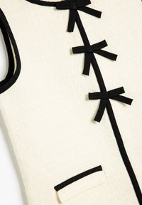 Abito in tessuto cremoso con dettagli neri, caratterizzato da una linea centrale di piccoli fiocchi neri e una tasca con bordo a contrasto.