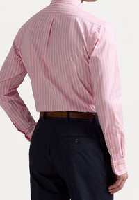 Růžová pruhovaná košile s dlouhými rukávy a zadním pásem, v kombinaci s tmavými kalhotami. Obsahuje manžety na knoflíky a klasický límeček.