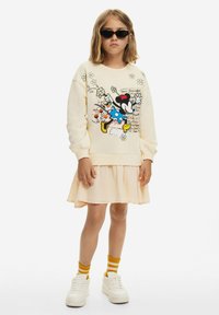 Felpa crema con una grafica colorata di un personaggio, abbinata a un vestito a strati rosa chiaro, sneakers bianche e calze a righe gialle.