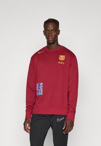 Μαρούν φούτερ με στρογγυλή λαιμόκοψη, κίτρινο λογότυπο Nike, έμβλημα της Μπαρσελόνα και μπλε σχέδιο στην τσέπη. Συνδυασμένο με μαύρο παντελόνι.