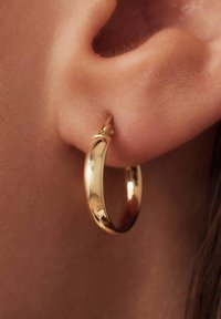 Boucle d'oreille en or suspendue à une oreille. La boucle d'oreille a une surface lisse et brillante, avec une forme arrondie et un design minimaliste.