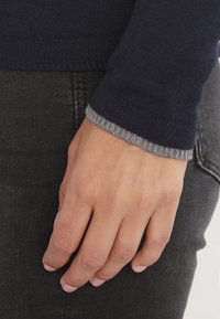 Marinblå stickad tröja med en ljusgrå ribbad accent, bärs tillsammans med mörka jeans. Närbild av en hand som vilar på denimtyget.